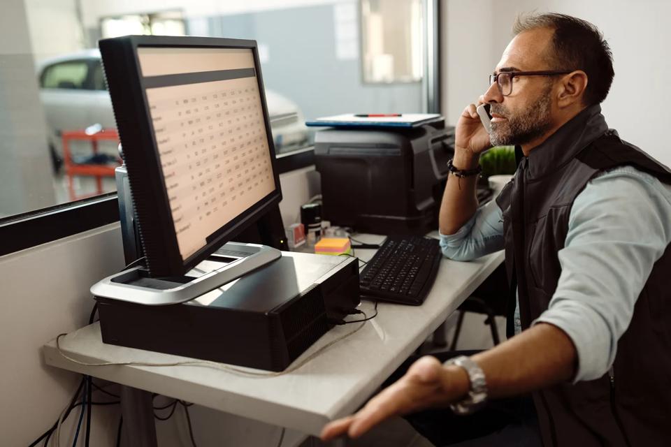 Foreman of auto mechanic workshop talking on the phone while using computer in the office.